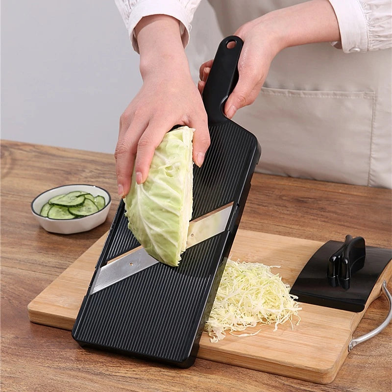 A hand is using a black vegetable grater tool to shred a piece of green cabbage on a wooden cutting board.