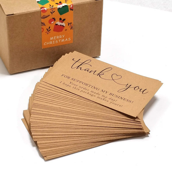 Ein Stapel brauner Kartenspiel-Dankesbriefe neben einer braunen Geschenkbox mit Weihnachtstags-Anhänger.