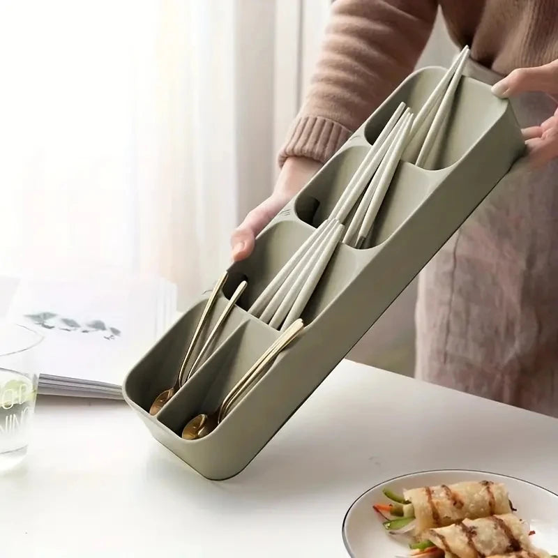 Una persona sostiene una bandeja organizadora de cubiertos rectangular, verde oliva y compartimentada sobre una mesa blanca.