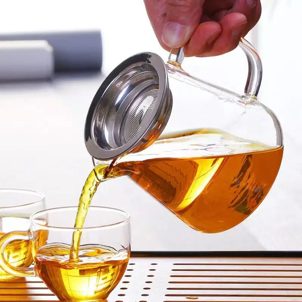 Eine Hand gießt bernsteinfarmenten Tee aus einem silbernen Teekessel in eine Glastasse.