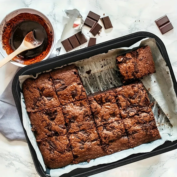 Dunkler Schokoladenbrownie in einer schwarzen Antihaft-Backform mit losem Schokoriechen dazu.