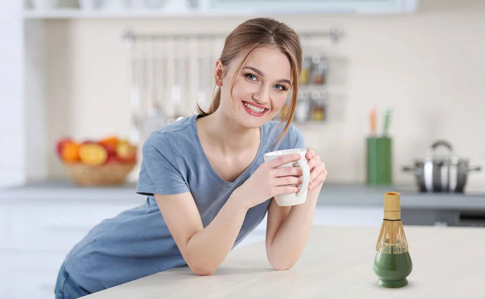 A smiling woman holds a white ceramic matcha whisk in a bright kitchen setting.