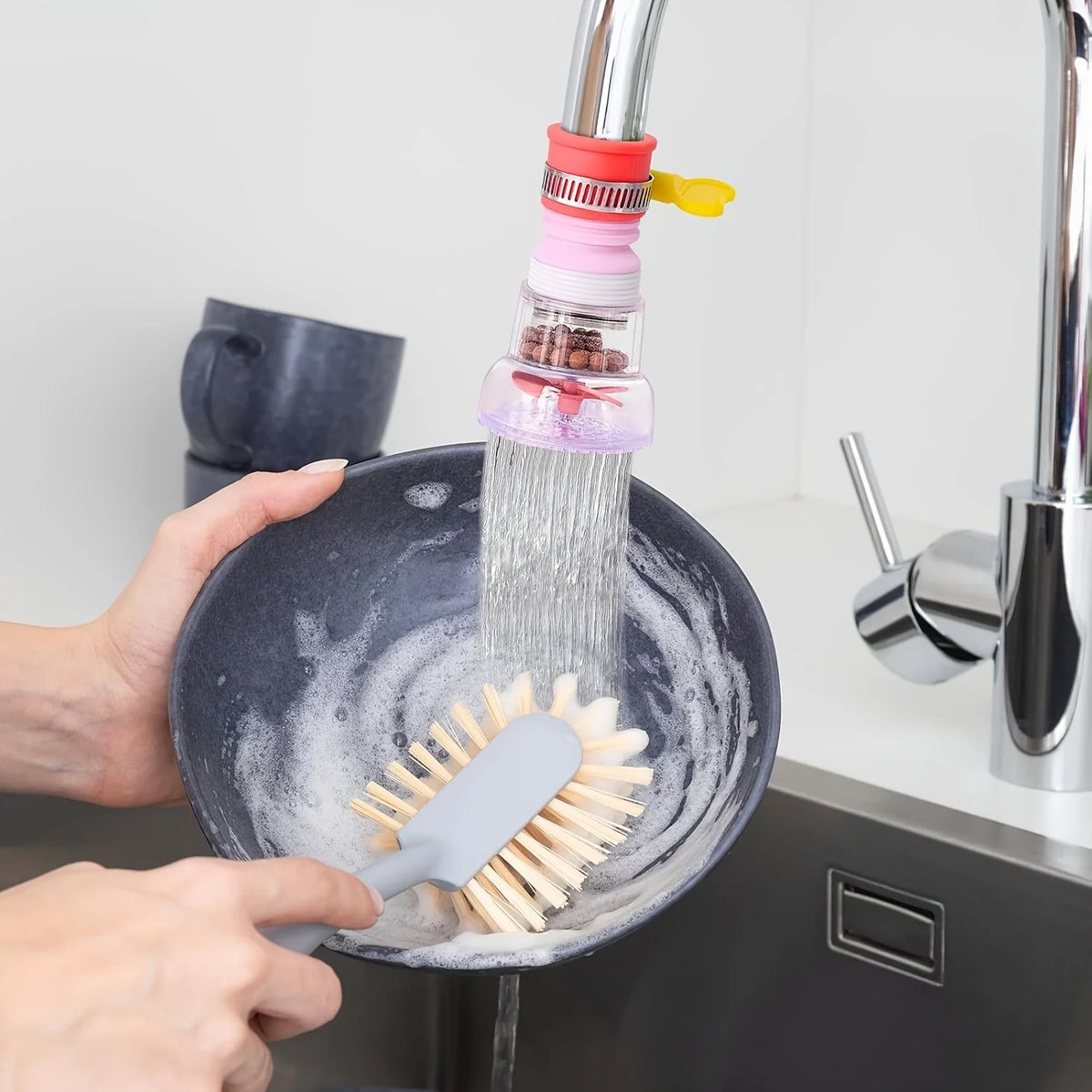 Manos usando un extensor de grifo y cepillo de cocina para limpiar un plato bajo un chorro de agua.