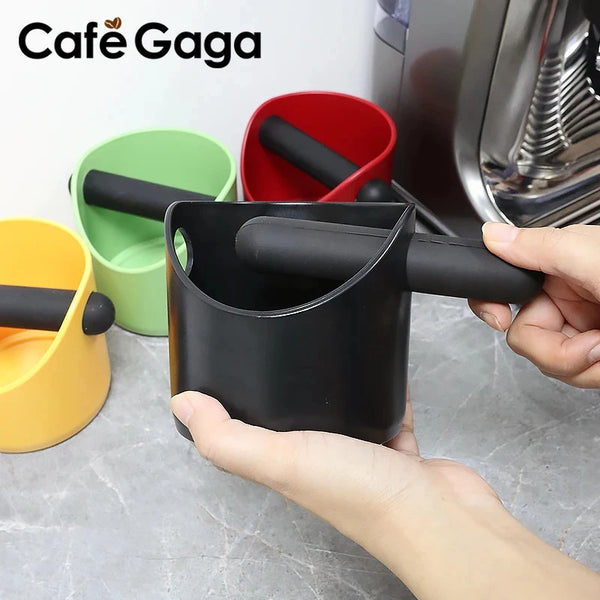 Eine Hand hält eine schwarze Kaffeeknopf-Box mit einem schwarzen Kolben neben farbigen Tassen.