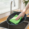 A hand is wiping a black frying pan with a bright green microfiber cleaning cloth in a kitchen setting.