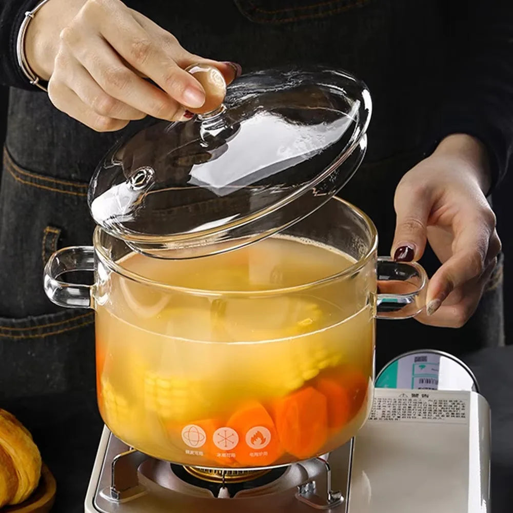 A hand lifts the lid of a glass stew pot filled with yellowish liquid and vegetables on a heating plate.
