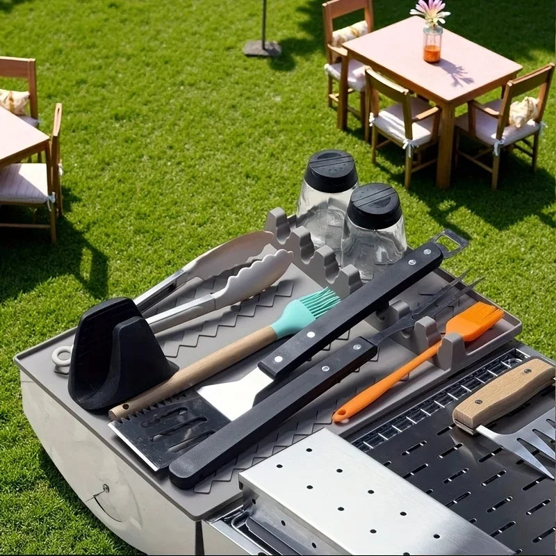 A silicone side burner rack and utensil storage mat with various kitchen tools placed on a metal surface outdoors.