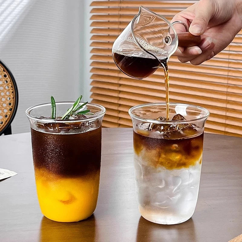 A hand pours dark liquid from a glass into a clear tumbler filled with ice and amber-colored liquid.