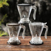 Clear glass pour over coffee drippers with handles resting on a wooden surface.