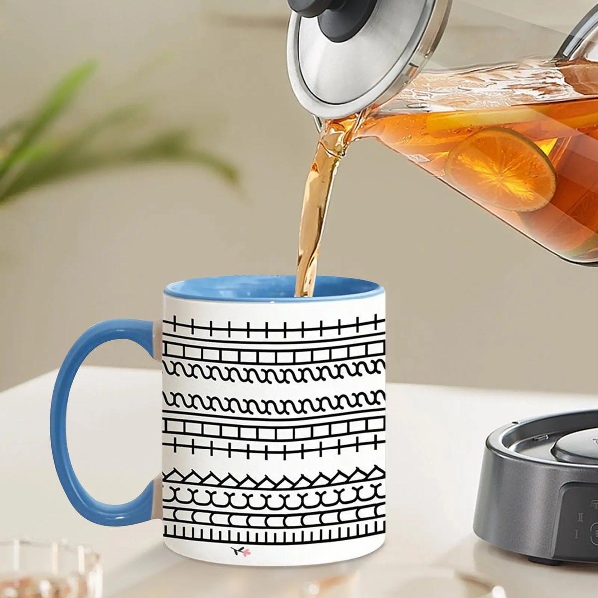 A white ceramic coffee mug with black decorative patterns is being filled with orange juice from a glass pitcher.