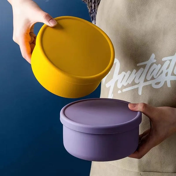 Eine Hand hält eine gelbe Silikon-Lunchbox und einen lila Silikonbehälter vor einem dunkelblauen Hintergrund.