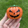 An orange pumpkin-shaped manual herb and spice grinder with a green lid being held in a hand against a green background.