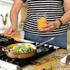 A person holds an airtight glass storage jar with a bamboo lid over a sizzling pan on a stovetop.
