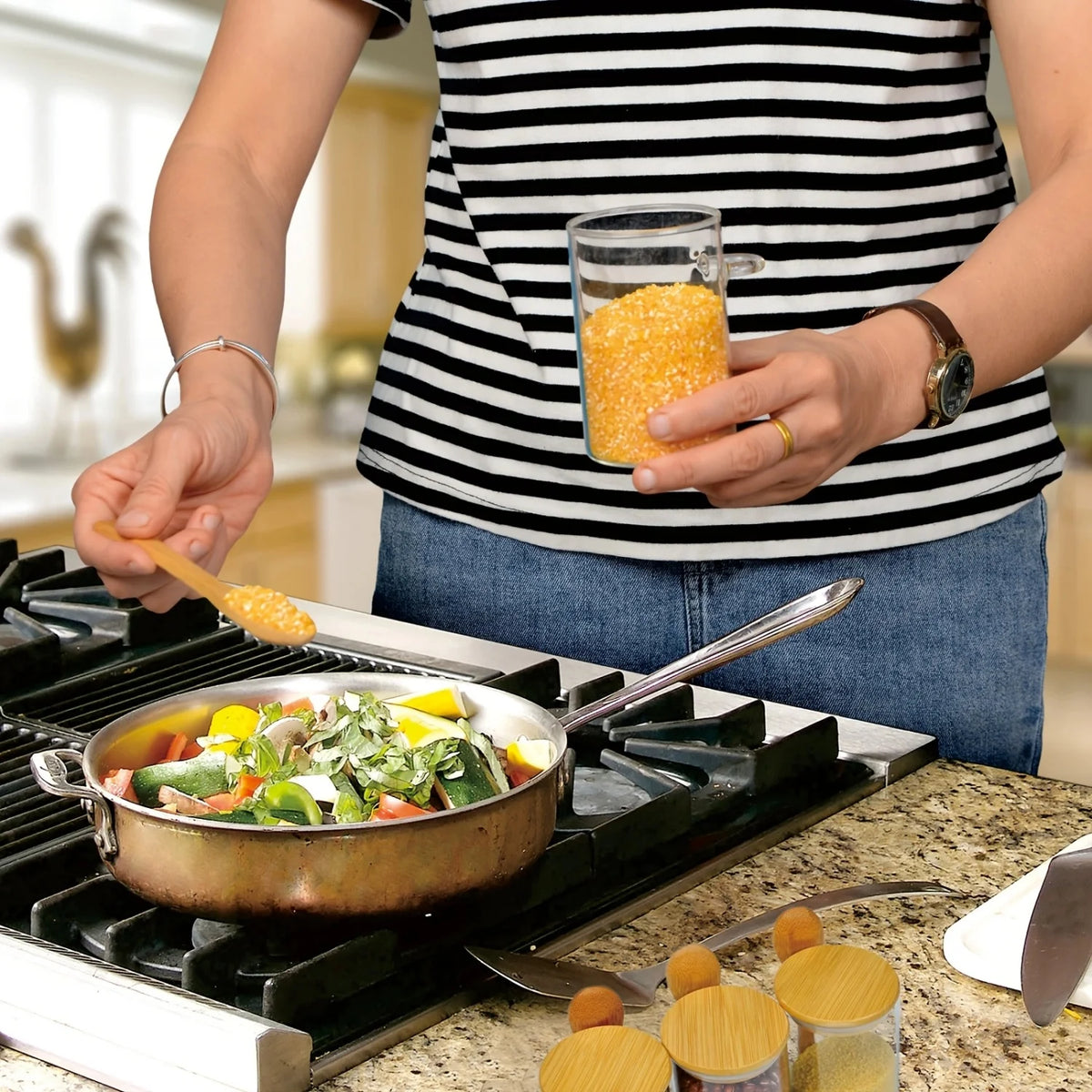 A person holds an airtight glass storage jar with a bamboo lid over a sizzling pan on a stovetop.