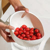 A hand is pouring red tomatoes through a white multi-purpose kitchen strainer and sieve.