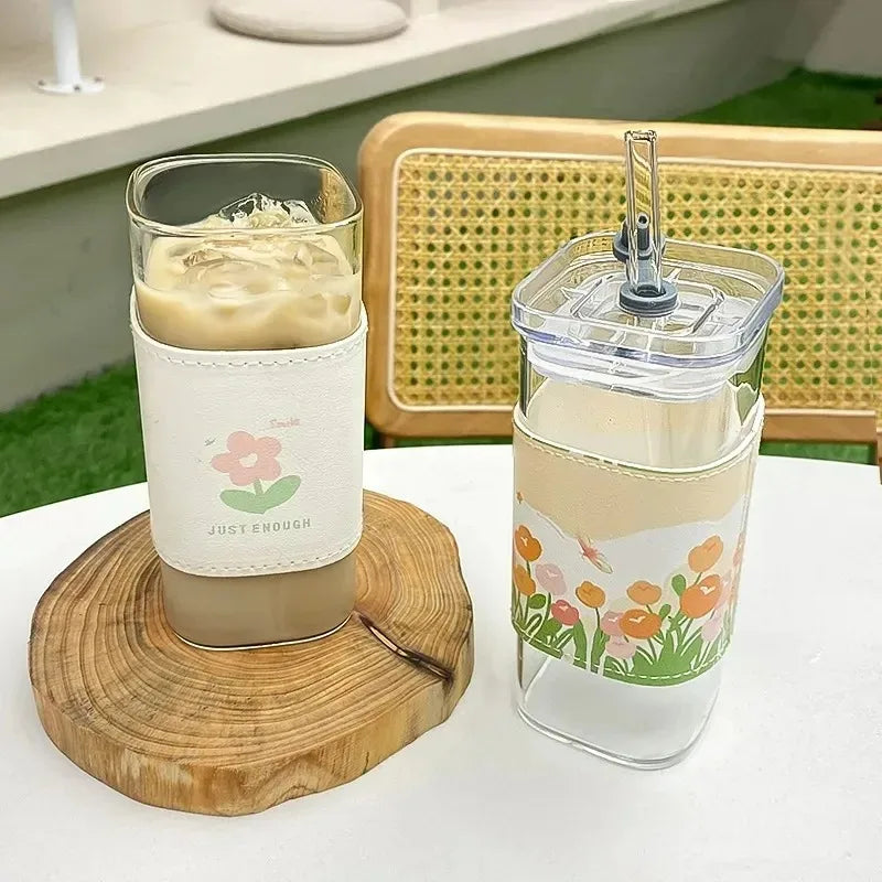 A square heat resistant glass drink cup with a lid and straw filled with a light beige beverage sits on a wooden coaster.