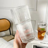 A hand holds a clear drinking glass with a lid and straw, showing its ribbed texture.