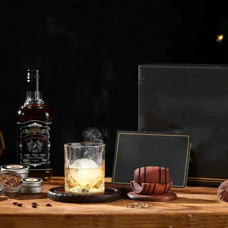A glass of whiskey is shown next to a dark wooden infuser kit containing wood chips and spices on a wooden surface.