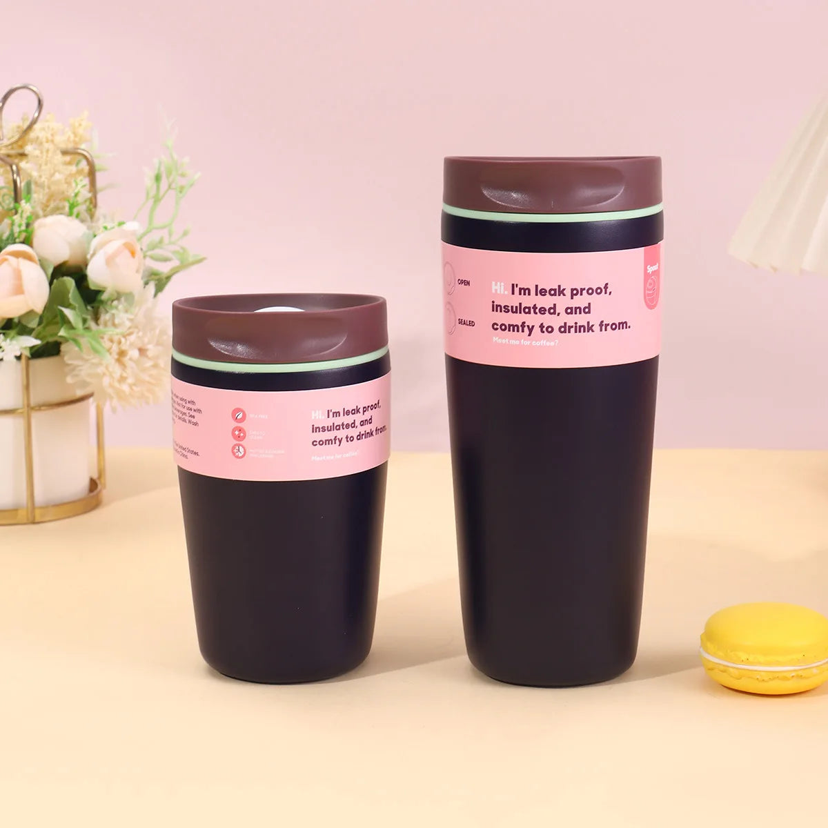 A dark brown vacuum insulated coffee mug with a pink label sits on a light surface next to a yellow pastry.