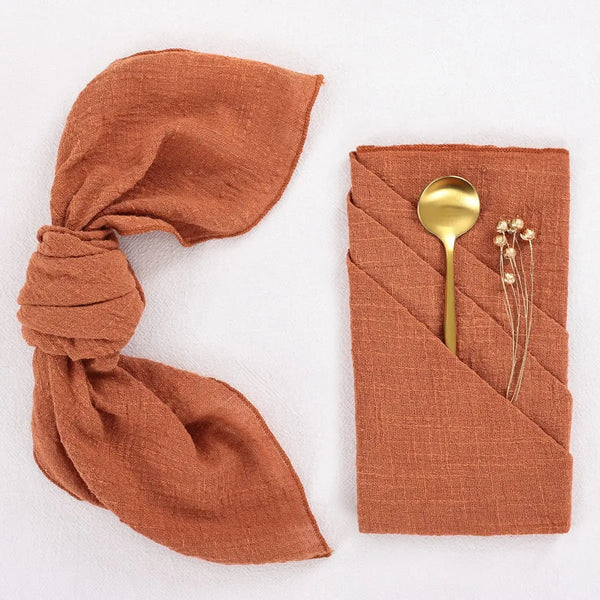 Ein Set aus drei rostfarbenen, natürlichen Mullservietten, eine mit Schleife gebunden und eine mit goldener Gabel gefaltet.
