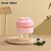 A small pink reusable food container with a clear lid is shown resting on a woven mat next to a blueberry.
