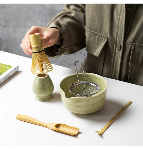 Eine Hand gießt Matcha-Pulver aus einem Bambusbesen in eine hellgrüne Keramikschale, mit einem Holzlöffel daneben.