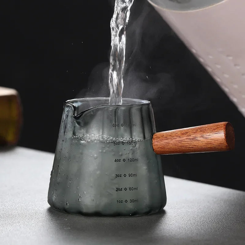 Una taza medidora de espresso de vidrio con asa de madera se está llenando de agua, mostrando marcas de volumen precisas.