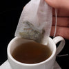 A hand holds a white disposable non-woven tea filter bag filled with dried green tea leaves above a white cup of brewed tea.
