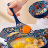 A hand is using a blue ceramic serving spoon to scoop food from a bowl of soup containing corn and other ingredients.