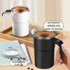 A hand holds a black automatic stirring coffee cup on a wooden tray with a cup lid and coffee beans.