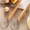 A stainless steel and wood dough whisk set with a wire whisk and a balloon whisk resting on a wooden surface next to eggs.