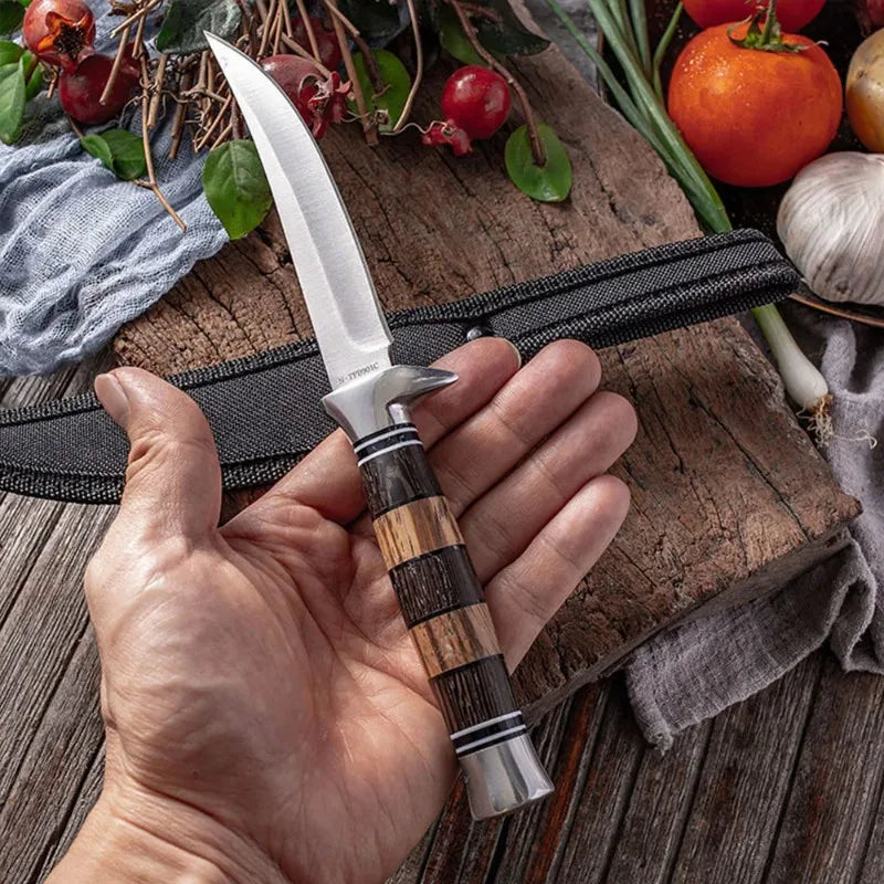 A hand holds a Japanese style sashimi knife with a wooden handle resting on a rustic cutting board surrounded by fresh vegeta