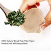A hand holds a wooden tea scoop filled with green tea leaves and a natural wood pulp filter paper bag.