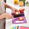 A hand is using a black knife to cut a tomato on a purple plastic cutting board, with fresh vegetables nearby.