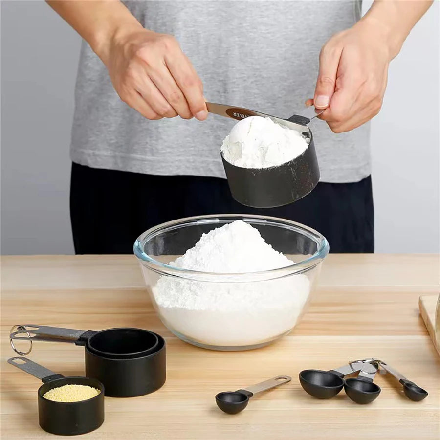 Hands are measuring white powder into a glass bowl using a stainless steel measuring cup and spoon set for baking.