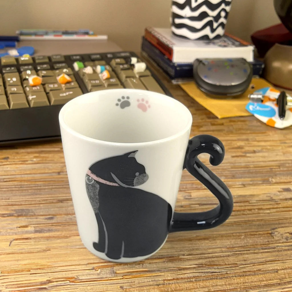 Weiße Keramiktasse mit schwarzen Katzen-Silhouette und Schwanzhenkel auf Holztable