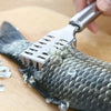 A stainless steel fish scale remover tool is shown being used on a textured, dark fish skin on a wooden surface.