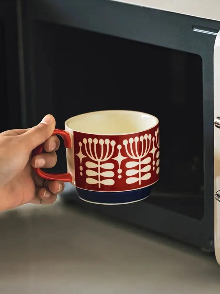Une main tient une tasse à café en céramique rouge et blanc devant un appareil sombre.
