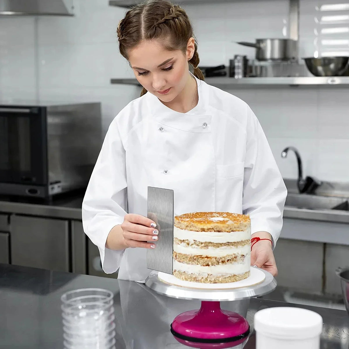 A woman is decorating a layered cake with a stainless steel scraper set in a kitchen environment.