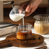 A hand pours milk from a glass pitcher into a glass milk pot for tea or espresso.