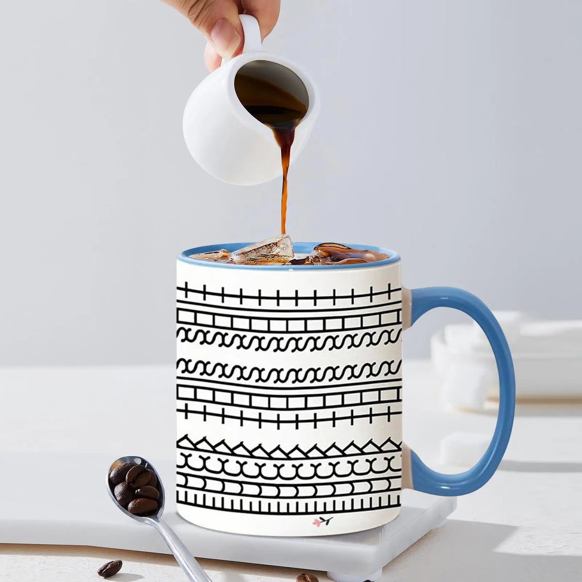 A hand pours dark coffee from a white cup into a blue ceramic mug decorated with black geometric patterns.