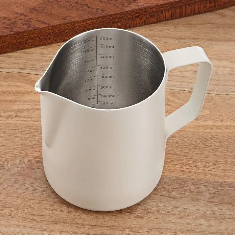 A stainless steel coffee frothing pitcher with various volume markings is shown on a wooden surface.