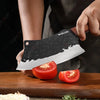 A hand holds a black forged chef knife with a white blade, positioned above a wooden cutting board.