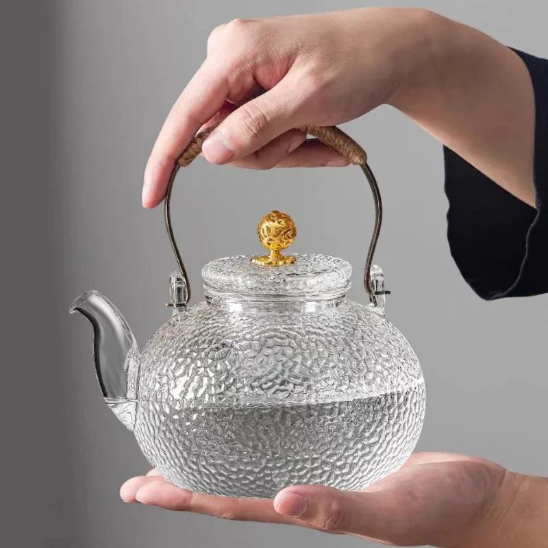 A hand holds a hand-hammered glass teapot with a gold lid and hemp rope handle.
