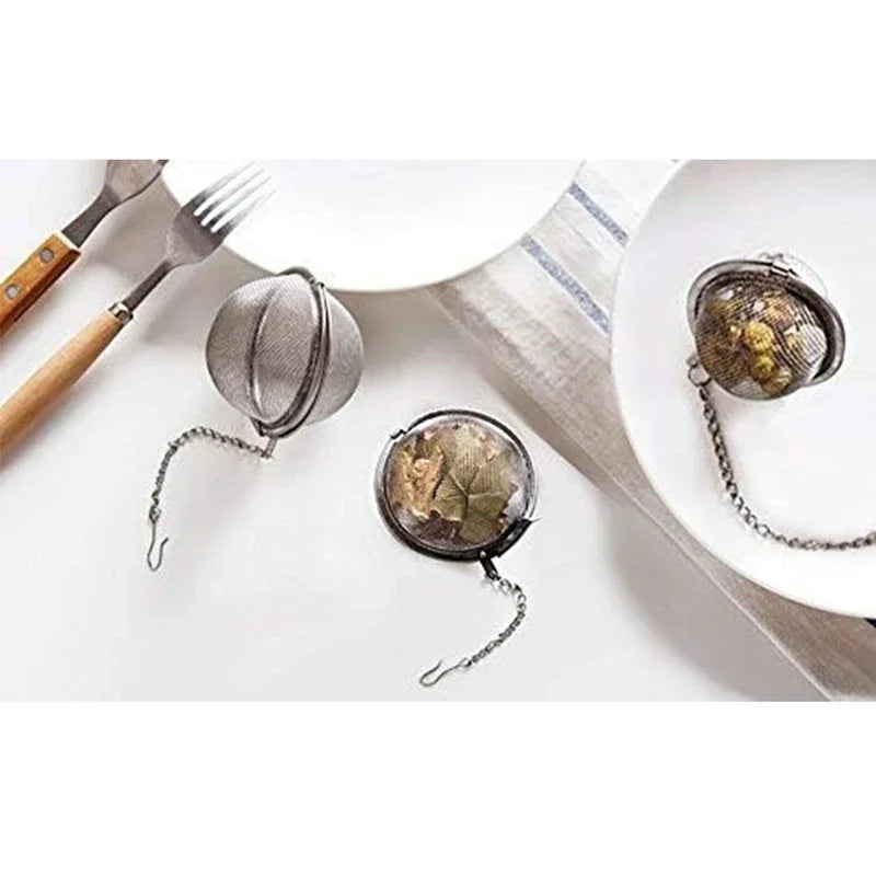 Stainless steel mesh tea infuser with hook resting on a white plate next to a fork.