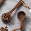 A wooden coffee measuring spoon with a long handle and a small wooden scoop filled with roasted coffee beans on a gray surfac