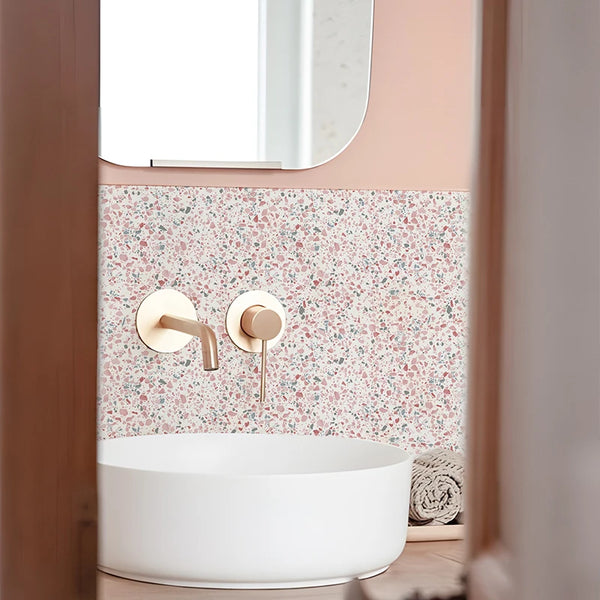 A white vessel sink is mounted on a pink terrazzo wall with brass fixtures and a mirror above.