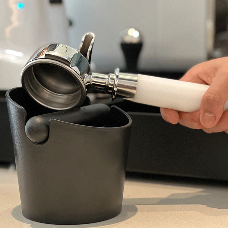 A hand holds a silver espresso knock box with a white handle, positioned above a dark container.