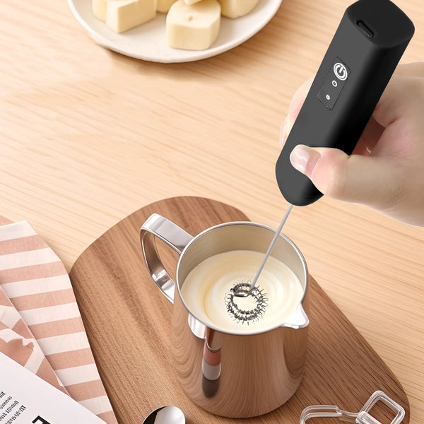 A hand pours milk into a stainless steel pitcher using a portable electric milk frother and blender.