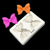 Three silicone baking molds shaped like bows in orange, pink, and white are displayed on a white square surface.